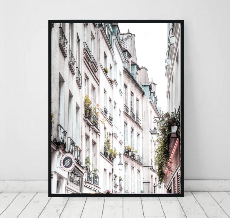 May include: A narrow street in Paris, France, with tall white buildings and balconies. The buildings have windows and doors, and some have plants growing on them. The street is lined with trees and there is a lamppost in the middle of the street.