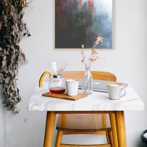 May include: A white marble table with a wooden chair and a glass coffee pot with a wooden handle. There is a white ceramic mug and a laptop on the table. A framed print of a foggy forest scene hangs on the wall behind the table.