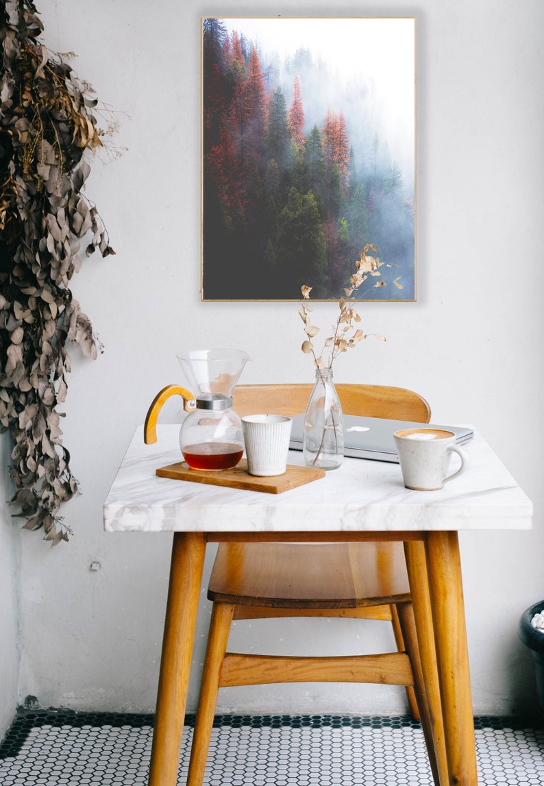 May include: A white marble table with a wooden chair and a glass coffee pot with a wooden handle. There is a white ceramic mug and a laptop on the table. A framed print of a foggy forest scene hangs on the wall behind the table.