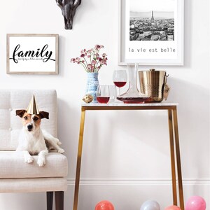 May include: A gold metal console table with a white marble top. The table is decorated with a bouquet of pink flowers in a blue vase, two wine glasses, a decanter, and a gold ice bucket. A dog wearing a party hat sits on a white chair in the background. The text "la vie est belle" is printed on a black and white framed photo hanging on the wall.