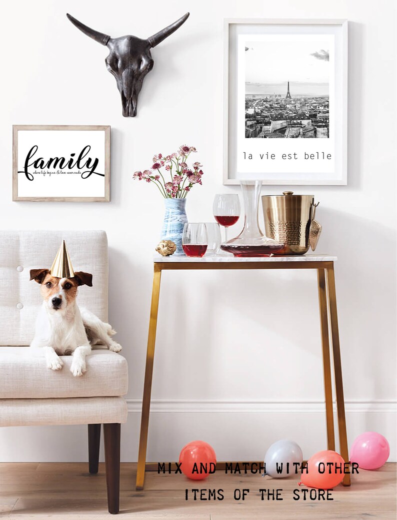 May include: A gold metal console table with a white marble top. The table is decorated with a bouquet of pink flowers in a blue vase, two wine glasses, a decanter, and a gold ice bucket. A dog wearing a party hat sits on a white chair in the background. The text "la vie est belle" is printed on a black and white framed photo hanging on the wall.