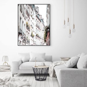 May include: A photograph of a narrow street in Paris, France. The street is lined with tall, white buildings with balconies and windows. The buildings are in a row, and the street is narrow, with a few people walking on the sidewalk.