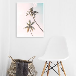 May include: A framed print of two palm trees against a pink and blue sky. The print is hanging on a white wall above a white chair with wooden legs.
