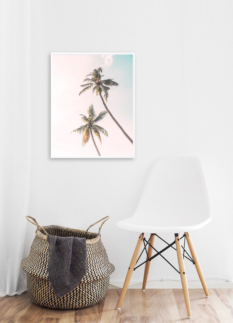 May include: A framed print of two palm trees against a pink and blue sky. The print is hanging on a white wall above a white chair with wooden legs.