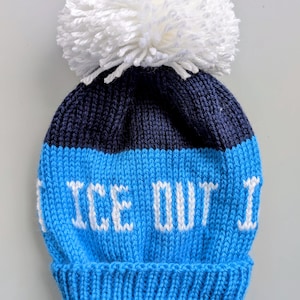 May include: A blue and navy knit beanie with a large white pom-pom. The beanie features the text "ICE OUT" in white block letters across the front. The hat is designed for cold weather.