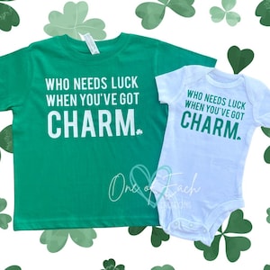 May include: A green t-shirt and a white onesie with the text "WHO NEEDS LUCK WHEN YOU'VE GOT CHARM." The design includes a small shamrock. The background features shamrock shapes.