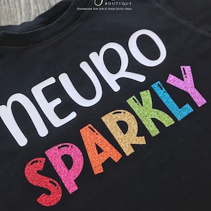 May include: Black t-shirt with the words "Neuro Sparkly" in white and rainbow glitter lettering.