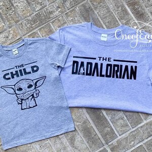 May include: Two gray t-shirts with black text and graphics. One shirt says "THE CHILD" with a cartoon character. The other says "THE DADALORIAN". Both shirts are short-sleeved and have a crew neck.