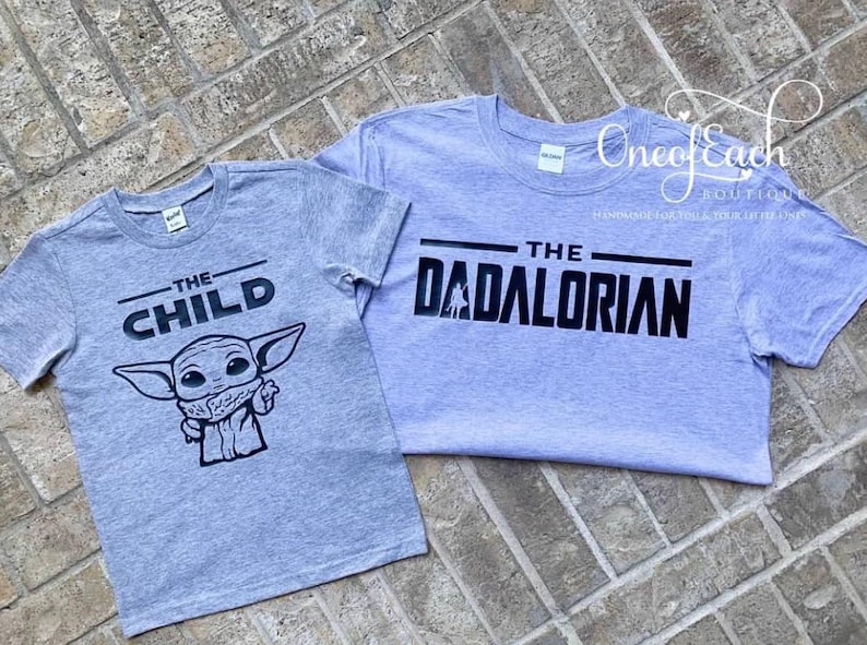 May include: Two gray t-shirts with black text and graphics. One shirt says "THE CHILD" with a cartoon character. The other says "THE DADALORIAN". Both shirts are short-sleeved and have a crew neck.