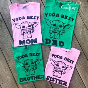 May include: Four t-shirts in pink and green, each with a black graphic of a character and the words "YODA BEST" above and "MOM", "DAD", "BROTHER", and "SISTER" below. The shirts are laid out on a wooden surface.