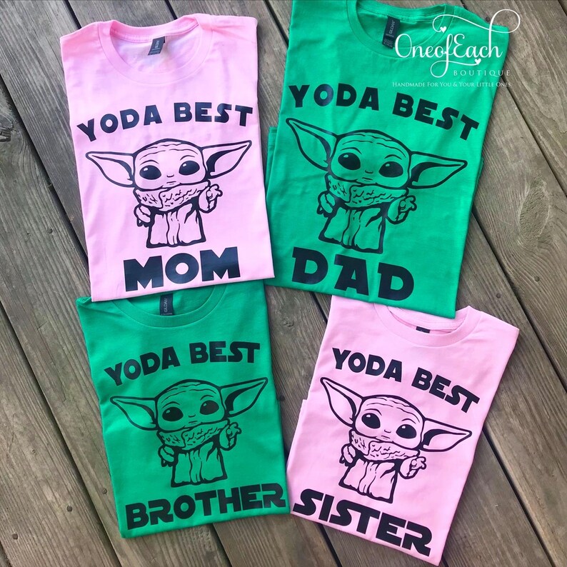 May include: Four t-shirts in pink and green, each with a black graphic of a character and the words "YODA BEST" above and "MOM", "DAD", "BROTHER", and "SISTER" below. The shirts are laid out on a wooden surface.