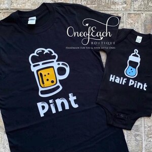May include: Black t-shirt with a white and yellow beer mug graphic and the word "Pint". A black baby onesie with a blue bottle graphic and the words "Half Pint". Both items are from One of Each Boutique.