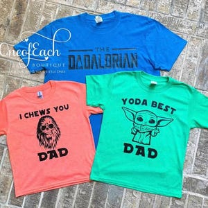 May include: Three colorful t-shirts with Star Wars-themed designs. One is blue with "The Dadalorian" text, another is coral with a Chewbacca graphic and "I Chews You Dad" text, and the third is green with a Yoda graphic and "Yoda Best Dad" text.