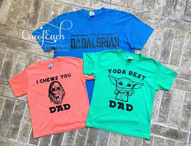 May include: Three colorful t-shirts with Star Wars-themed designs. One is blue with "The Dadalorian" text, another is coral with a Chewbacca graphic and "I Chews You Dad" text, and the third is green with a Yoda graphic and "Yoda Best Dad" text.