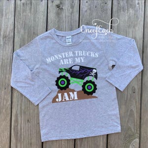 May include: A gray long-sleeved shirt featuring a graphic of a monster truck with the text "MONSTER TRUCKS ARE MY JAM". The monster truck is black with green and purple accents and a skull and crossbones.