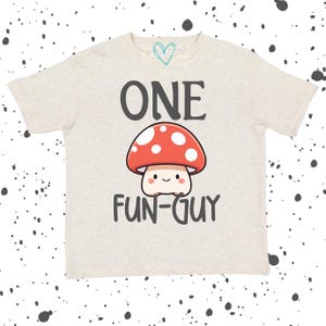 May include: A light beige t-shirt with the words "ONE FUN-GUY" in dark gray. A cartoon mushroom with a red cap and white spots is centered below the word "ONE". The shirt has short sleeves and a crew neck.