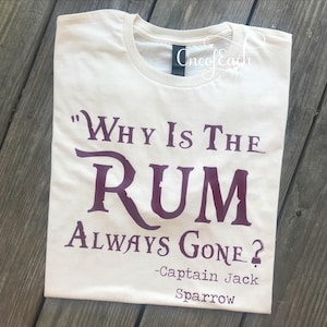 May include: Cream-colored t-shirt with the quote "Why Is The Rum Always Gone?" in dark purple, attributed to Captain Jack Sparrow. The shirt is folded and displayed on a wooden surface.