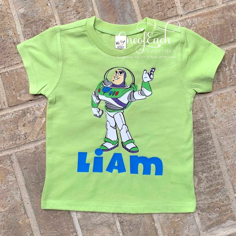 May include: Green short-sleeved t-shirt with a cartoon image of Buzz Lightyear from Toy Story. The shirt has the name "Liam" printed in blue letters below the image.