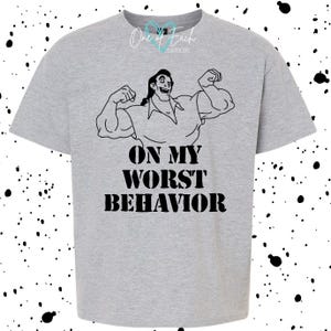 May include: A light gray t-shirt featuring a black illustration of a muscular man flexing his biceps. The text "ON MY WORST BEHAVIOR" is printed in bold, black letters below the illustration. The shirt has a crew neck.