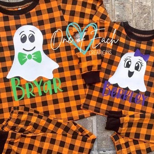 May include: Two long-sleeve pajama sets with an orange and black buffalo plaid pattern. Each shirt features a white ghost graphic with text reading "One of Each CREATIONS". One ghost has a green bow tie and the name "BRYAR", the other a purple bow and "BERKLEY".