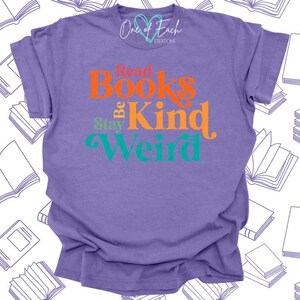 May include: A purple t-shirt with the words "Read Books Be Kind Weird" in orange, teal, and green. The shirt also has the text "One of Each Creations" in white. The shirt is made of a soft material.