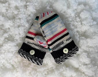 May include: A pair of knitted mittens with a colorful striped pattern in red, black, teal, and light blue. The cuffs are black with a white and black zig-zag design and a white button. The mittens are designed for cold weather.