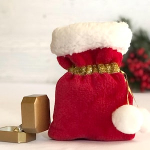 May include: A small red velvet drawstring pouch with a white faux fur trim at the top, a gold glittery drawstring, and two white pom-poms attached to the cord. Next to the pouch is an open, matte gold-colored hexagonal ring box.