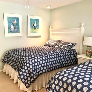 May include: A bedroom with two twin beds, each with a navy blue comforter featuring a white seashell pattern. White bed skirts and pillows with a similar design complement the bedding. Two framed watercolor paintings of birds hang on the wall.