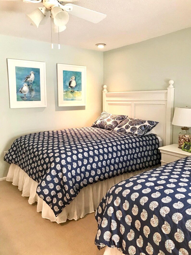 May include: A bedroom with two twin beds, each with a navy blue comforter featuring a white seashell pattern. White bed skirts and pillows with a similar design complement the bedding. Two framed watercolor paintings of birds hang on the wall.