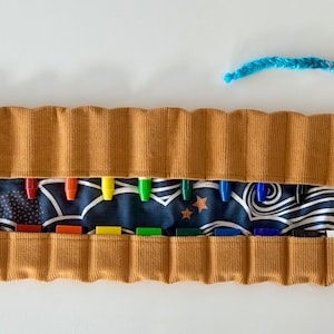 Könnte beinhalten: Eine braune Cord-Stifte-Rolle mit einer blauen und weißen gemusterten Innenausstattung. Die Rolle ist geöffnet und enthält eine Vielzahl von Buntstiften. Die Rolle hat eine blaue und braune geflochtene Schnur.