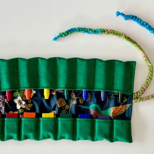 May include: A green corduroy pencil roll with a floral and hummingbird patterned interior. The roll is open, revealing colourful pencils and pens. A braided strap in shades of blue, green, and tan is attached.