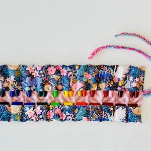 May include: A patterned crayon roll featuring a floral and swan design in blue, pink, and gold. The roll is open, displaying a selection of crayons. A braided cord in pink and blue is attached. The roll is open, displaying a selection of crayons.