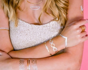 May include: A woman wearing a white sequined top with gold and silver temporary tattoos on her arms and a gold necklace with a silver pendant.