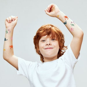 May include: A young person with red hair wearing a white t-shirt and temporary tattoos of sea creatures on their arms. The tattoos include a whale, a crab, a starfish, a seahorse, and an octopus.