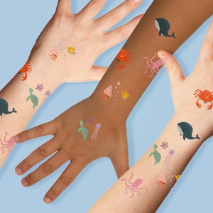May include: Three arms with temporary tattoos of various sea creatures, including a whale, octopus, turtle, crab, and seashells. The tattoos are in a variety of colors, including blue, green, pink, orange, and yellow.