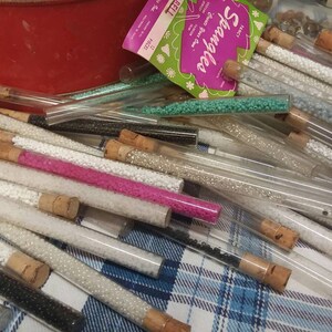 Op de afbeelding: Een verzameling glazen buizen gevuld met verschillende kleuren kleine kraaltjes. De buizen zijn met kurken afgesloten en gerangschikt op een geruite stof. Het etiket op een van de buizen luidt "Sparkle, Great Party Gift, 127", en "Price 44c".