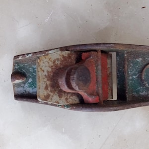 Puede incluir: Un cepillo de mano vintage con un cuerpo de metal verde y rojo. El cepillo tiene una cuchilla de metal y un mango de madera.