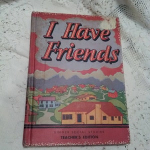 Pode incluir: Livro vintage vermelho e amarelo intitulado "I Have Friends" com uma imagem de uma pequena cidade na capa. O livro é rotulado como "Singer Social Studies Teacher's Edition".