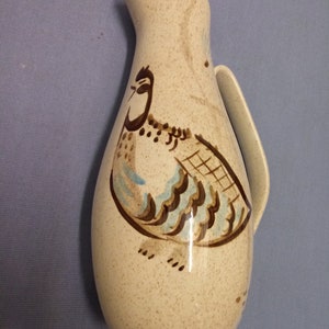 Puede incluir: Una jarra de cerámica beige con un diseño pintado de un pájaro marrón y azul. La jarra tiene una textura moteada y un asa.