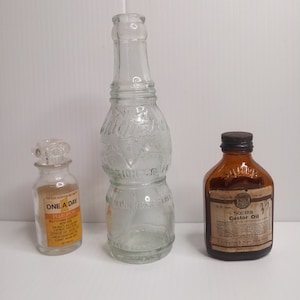 Peut inclure: Trois bouteilles en verre, une transparente avec une surface texturée, une transparente avec une étiquette qui indique "One A Day Plus Iron", et une bouteille ambrée avec une étiquette qui indique "Squibb Castor Oil".