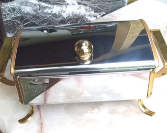 May include: A rectangular, stainless steel chafing dish with gold-colored accents. The lid has a gold knob, and the dish has gold handles and feet. The surface reflects the surroundings, creating a shiny appearance. This serving dish is suitable for buffet-style meals.