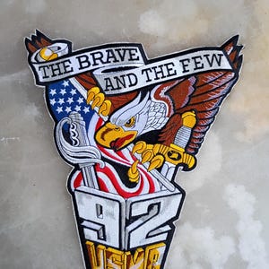 Puede incluir: Parche bordado con un águila sosteniendo una espada y una pancarta con el texto "THE BRAVE AND THE FEW". El diseño incorpora la bandera estadounidense, el número "92" y "USMA" en una combinación de colores patrióticos.