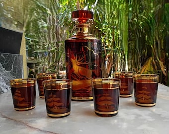 May include: A deep amber glass decanter with a matching stopper and five short tumblers. The decanter features an etched stag design, while each tumbler displays an etched pine tree motif. All pieces have gold-toned rims and bases.