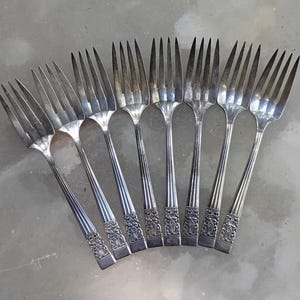 May include: Eight silver forks are fanned out, each with multiple tines and a long handle. The handles have vertical grooves and a decorative floral design near the base. The forks are displayed on a light grey surface.