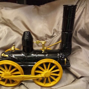 May include: A black and yellow model train with a tall smokestack and large yellow wheels. The train has gold-coloured accents. The train is a vintage style and is set against a neutral background.