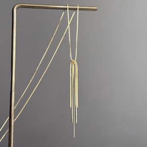 Puede incluir: Un collar de tono dorado con una cadena fina de eslabones cuadrados y un colgante compuesto por tres barras metálicas verticales en forma de U de diferentes longitudes, dispuestas en niveles para crear un diseño lineal, limpio y simétrico.