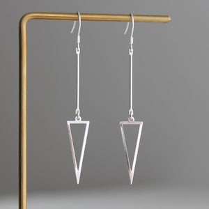 Può includere: Un paio di orecchini pendenti color argento caratterizzati da una lunga e sottile barra verticale collegata a un ciondolo triangolare cavo rivolto verso il basso. Gli orecchini sono sospesi a ganci standard dal design geometrico.