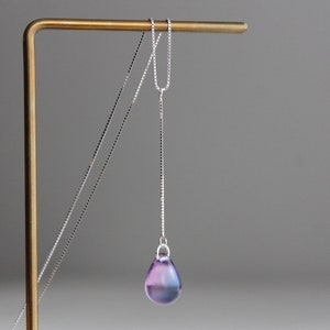 Può includere: Una collana a catena d'argento con un ciondolo a forma di lacrima. Il ciondolo è in vetro iridescente con sfumature di viola e blu.