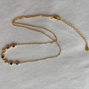 Puede incluir: Un collar dorado con una serie de piedras preciosas de colores. Las piedras están dispuestas en semicírculo, mostrando un espectro de colores. El collar tiene una cadena delicada y un cierre ajustable.