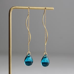 Puede incluir: Un par de pendientes de gota de oro con gotas de vidrio azul que cuelgan de una cadena curva.